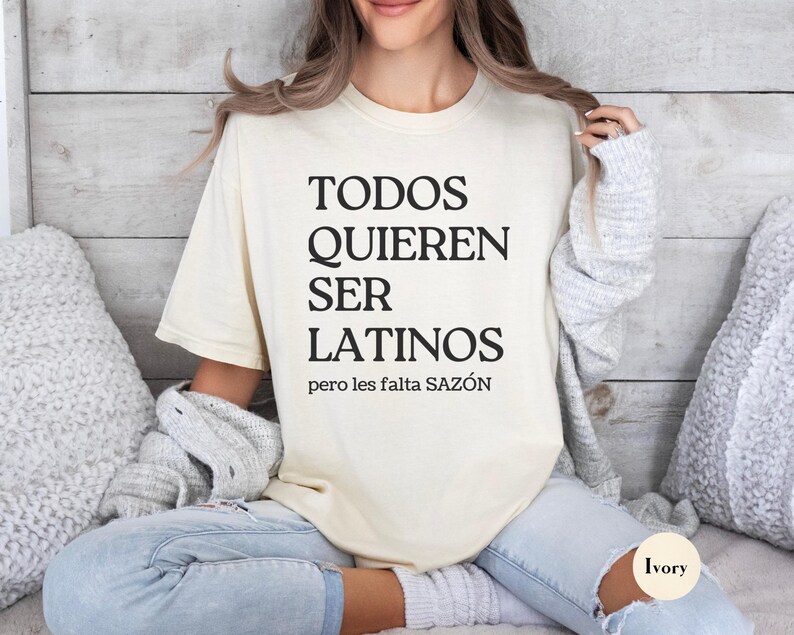 May include: Ivory-colored t-shirt with the text "TODOS QUIEREN SER LATINOS pero les falta SAZ&Oacute;N" in black. The shirt is a relaxed fit, with a crew neck. The model is wearing light-wash jeans.