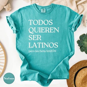May include: Seafoam green t-shirt with the text "TODOS QUIEREN SER LATINOS pero les falta SAZ&Oacute;N" in white. The shirt is folded, with rolled-up sleeves. A straw hat and succulent are in the background.