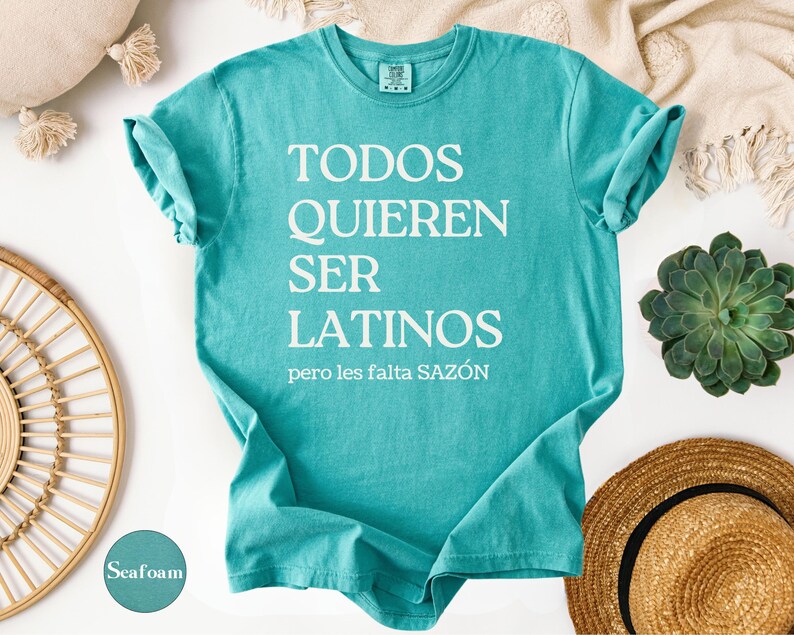 May include: Seafoam green t-shirt with the text "TODOS QUIEREN SER LATINOS pero les falta SAZ&Oacute;N" in white. The shirt is folded, with rolled-up sleeves. A straw hat and succulent are in the background.