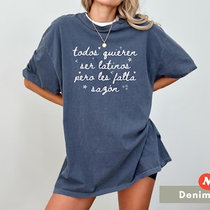 May include: A denim blue t-shirt with the text "todos quieren ser latinos pero les falta saz&oacute;n" in white script. The shirt has short sleeves and a relaxed fit. A small "New!" sticker is in the bottom right corner.