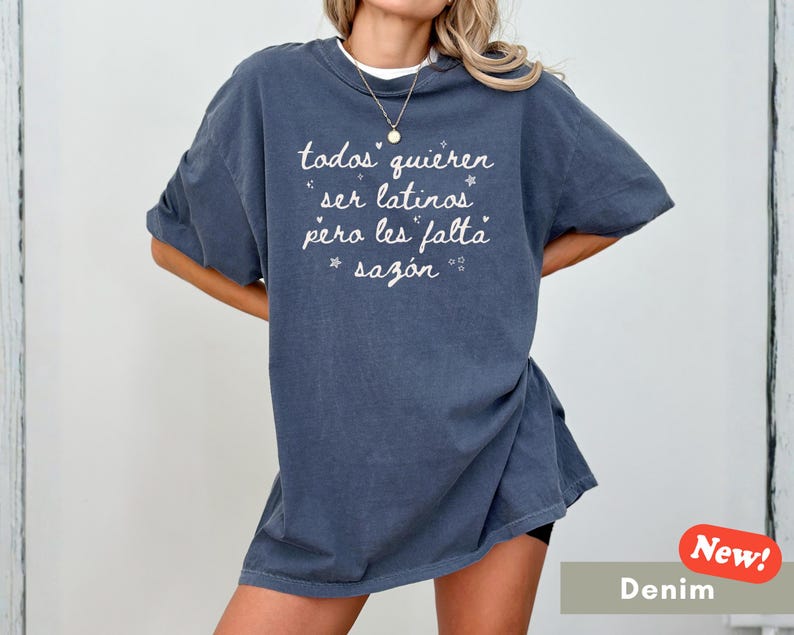 May include: A denim blue t-shirt with the text "todos quieren ser latinos pero les falta saz&oacute;n" in white script. The shirt has short sleeves and a relaxed fit. A small "New!" sticker is in the bottom right corner.