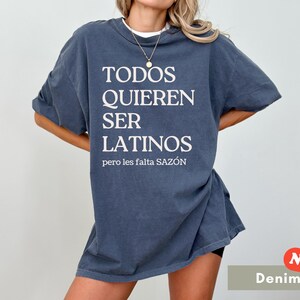 May include: Denim blue oversized t-shirt with the text "TODOS QUIEREN SER LATINOS pero les falta SAZ&Oacute;N" in white. The shirt has short sleeves and a crew neck. A "New!" sticker is in the bottom right corner.