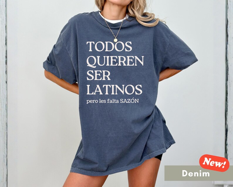 May include: Denim blue oversized t-shirt with the text "TODOS QUIEREN SER LATINOS pero les falta SAZ&Oacute;N" in white. The shirt has short sleeves and a crew neck. A "New!" sticker is in the bottom right corner.
