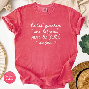 May include: A watermelon-colored t-shirt with rolled-up sleeves. The shirt features white script that reads "todos quieren ser latinos pero les falta saz&oacute;n." A small watermelon-colored circle with the word "Watermelon" is in the lower left corner.