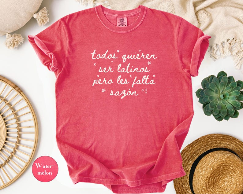 May include: A watermelon-colored t-shirt with rolled-up sleeves. The shirt features white script that reads "todos quieren ser latinos pero les falta saz&oacute;n." A small watermelon-colored circle with the word "Watermelon" is in the lower left corner.