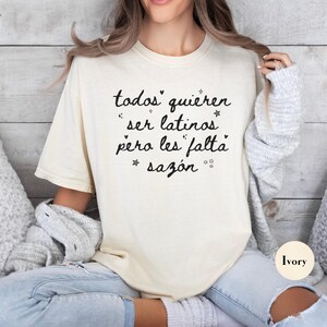 May include: Ivory-colored t-shirt with the text "todos quieren ser latinos pero les falta saz&oacute;n" in black script. The shirt is a relaxed fit and is paired with light-wash jeans. The background is a light wood panel.