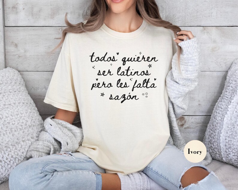 May include: Ivory-colored t-shirt with the text "todos quieren ser latinos pero les falta saz&oacute;n" in black script. The shirt is a relaxed fit and is paired with light-wash jeans. The background is a light wood panel.