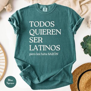 May include: Blue spruce t-shirt with the text "TODOS QUIEREN SER LATINOS" in white, with the phrase "pero les falta SAZ&Oacute;N" below. The shirt is folded, with a round "Blue Spruce" color swatch in the lower left corner.