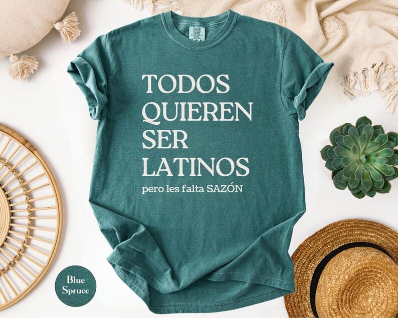 May include: Blue spruce t-shirt with the text "TODOS QUIEREN SER LATINOS" in white, with the phrase "pero les falta SAZ&Oacute;N" below. The shirt is folded, with a round "Blue Spruce" color swatch in the lower left corner.