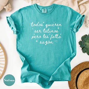 May include: Seafoam green t-shirt with rolled-up sleeves. The shirt features white text that reads "todos quieren ser latinos pero les falta saz&oacute;n" with star accents. The shirt is displayed on a white surface with decorative items.