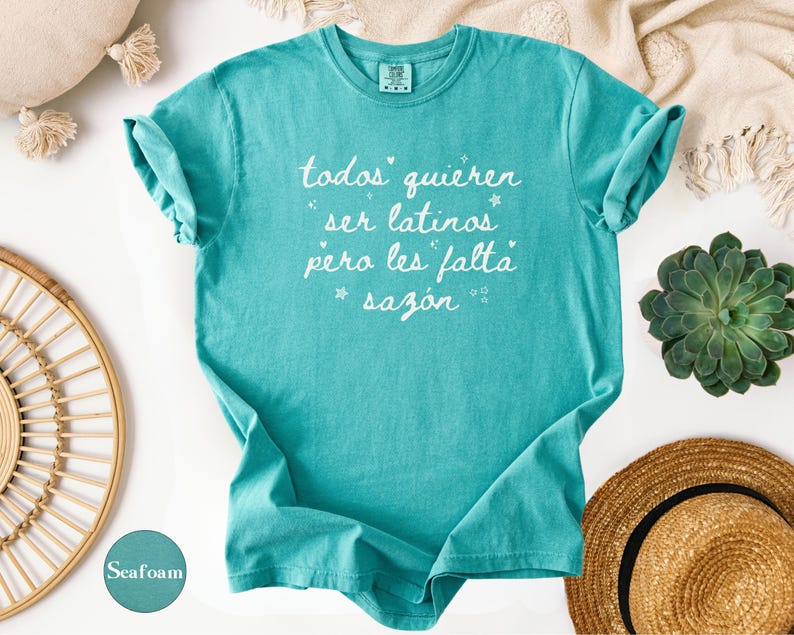 May include: Seafoam green t-shirt with rolled-up sleeves. The shirt features white text that reads "todos quieren ser latinos pero les falta saz&oacute;n" with star accents. The shirt is displayed on a white surface with decorative items.