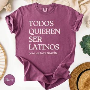 May include: Berry-colored t-shirt with the text "TODOS QUIEREN SER LATINOS pero les falta SAZ&Oacute;N" in white. The shirt is a crew neck style and is folded. A straw hat and a succulent plant are in the background.