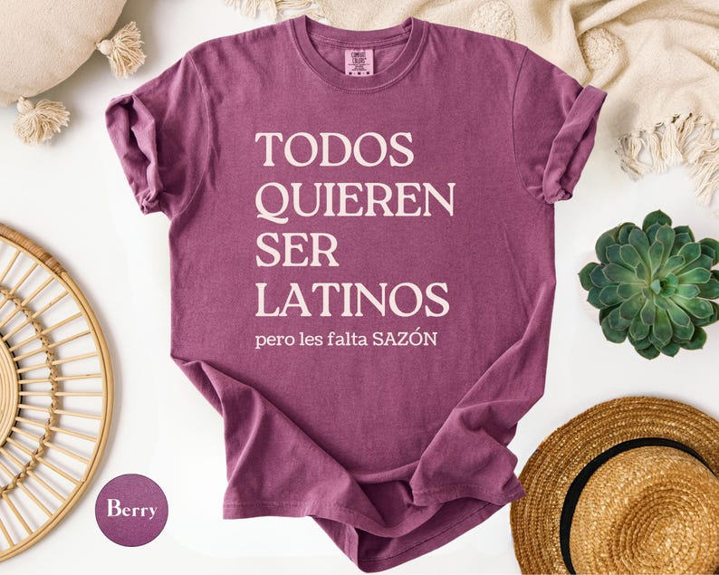 May include: Berry-colored t-shirt with the text "TODOS QUIEREN SER LATINOS pero les falta SAZ&Oacute;N" in white. The shirt is a crew neck style and is folded. A straw hat and a succulent plant are in the background.