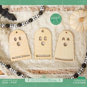Puede incluir: Tres recortes de fantasmas de madera con rasgos faciales circulares y ovalados, perfectos para manualidades de Halloween. Los recortes son de color marrón claro y tienen un borde inferior festoneado. Una guirnalda de cuentas en blanco y negro y una pequeña calabaza también están en la imagen. El texto "ready to CUT" es visible.
