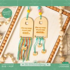 May include: Two wooden cat-themed ornaments with the text "On my way to pet my cat" and "On my way to pet my cats." Each has colourful yarn and cat-shaped tags with names. The background includes decorative wooden beads.