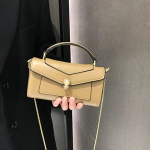 May include: Tan leather handbag with a gold chain strap and a top handle. The bag features a snake head clasp and a rectangular shape. The bag is held by a person wearing a black blazer.