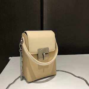 Puede incluir: Un pequeño bolso de mano color crema con una correa de cadena plateada. El bolso tiene forma rectangular con cierre de solapa y un cierre plateado. El cuerpo del bolso tiene una textura tejida y la solapa superior es lisa.
