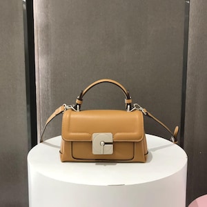 May include: A tan leather handbag with a top handle and a detachable shoulder strap. The bag has a rectangular shape with a flap closure and a silver-coloured clasp. The bag is on a white surface.