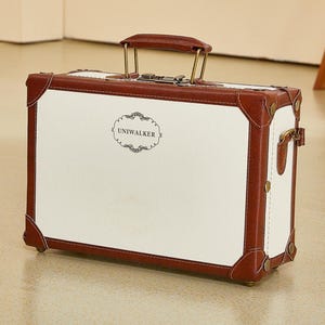 Op de afbeelding: Een vintage-stijl koffer met een witte body en bruine leren bekleding. De koffer heeft een handvat, metalen hardware en de tekst "UNIWALKER" op de voorkant. De hoeken zijn versterkt met bruin leer.