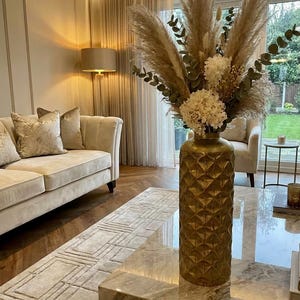 May include: A decorative gold vase with a geometric pattern, holding dried pampas grass, eucalyptus, and white flowers. The vase is on a marble coffee table in a living room with a beige sofa, rug, and curtains. The room has a warm, inviting atmosphere.
