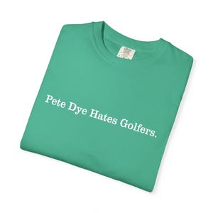 May include: A folded, teal-colored t-shirt with the text "Pete Dye Hates Golfers" printed in white. The shirt has a crew neck and a tag on the inside collar.
