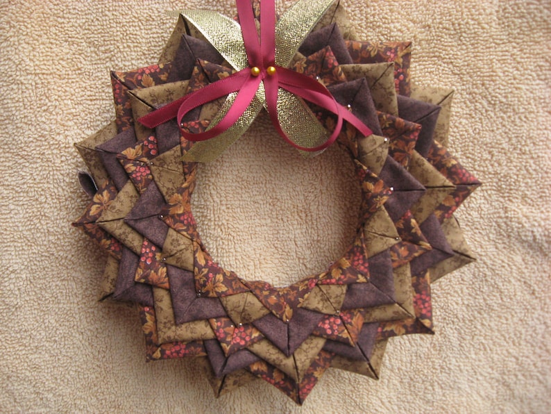 May include: A brown and gold fabric wreath with a red and gold bow in the center. The wreath is made of fabric squares sewn together in a star shape. The wreath is decorated with small red berries and gold glitter.