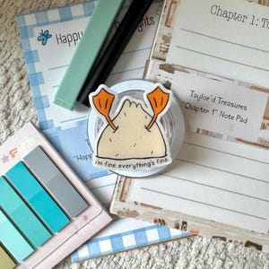 May include: A circular sticker featuring a cartoon beige dumpling with orange duck feet and the text 'i'm fine. everything's fine.' placed on a collection of stationery items including a blue checkered notepad and a set of blue sticky tabs.