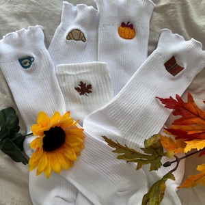May include: A collection of white ribbed crew socks featuring small embroidered designs including a blue coffee cup, a tan croissant, an orange pumpkin, a brown maple leaf, and an open brown book. The socks have ruffled top edges.