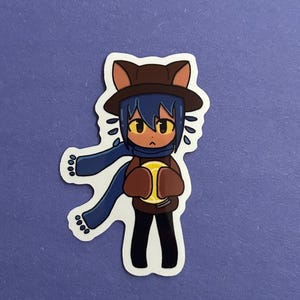 May include: A sticker depicting a cartoon character with cat-like features. The character is wearing a brown hat, blue scarf, and brown sweater, holding a yellow object. The sticker has a white border on a purple background.