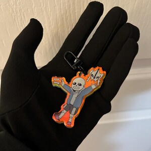 K&ouml;nnte beinhalten: Ein farbenfroher Acryl-Schl&uuml;sselanh&auml;nger mit einer Cartoonfigur in einer blauen Jacke, grauen Shorts und orangefarbenen Schuhen. Der Schl&uuml;sselanh&auml;nger hat einen schwarzen Verschluss und einen leuchtend orange-gelben Hintergrund.