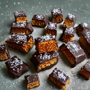 May include: A collection of rectangular honeycomb toffee pieces coated in dark chocolate and topped with coarse sea salt crystals. The interior of each piece reveals a porous, golden-brown honeycomb structure, while the exterior is smooth.