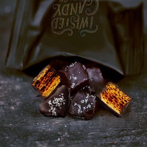 Pode incluir: Pedaços de doce de favo de mel cobertos com chocolate amargo e cristais de sal marinho, saindo de uma embalagem preta com o rótulo TWISTED CANDY. O interior dos pedaços de favo de mel apresenta uma textura porosa de cor castanho-dourada.