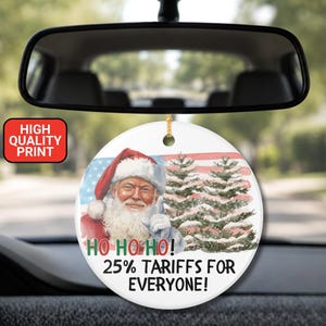 May include: A round white ceramic car ornament hanging from a rearview mirror. It features an illustration of Santa Claus, snow-covered pine trees, and a US flag background. Text reads: HO HO HO! 25% TARIFFS FOR EVERYONE! and a red box says HIGH QUALITY PRINT.