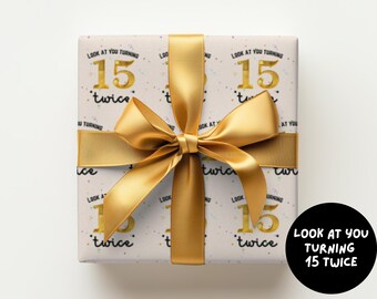 May include: A square gift box wrapped in white paper featuring a repeating pattern of the gold text 15 twice and the black text Look at you turning 15 twice, tied with a wide, shiny gold satin ribbon centered in a large, decorative bow.