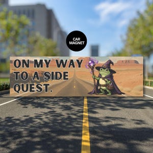 May include: A rectangular car magnet with the text "ON MY WAY TO A SIDE QUEST." A cartoon frog wizard with a staff and cape stands on a road leading to a desert landscape. A black circle above reads "CAR MAGNET."