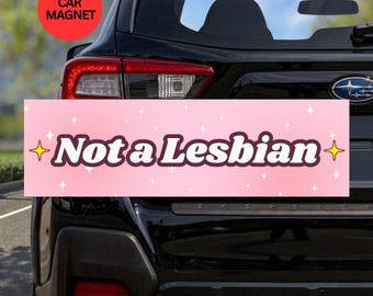 May include: A rectangular car magnet with a light pink background featuring white sparkles and yellow four-pointed stars. The text "Not a Lesbian" is printed in a bold, dark brown, rounded font across the center of the magnet.