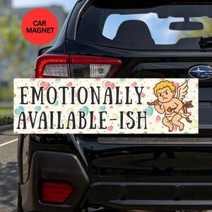 May include: A rectangular car magnet featuring a cream background with a pattern of small pastel hearts. The text reads EMOTIONALLY AVAILABLE-ISH in black capital letters. A cartoon cupid with blonde hair holds a bow and arrow on the right side.