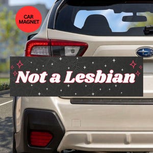 May include: A rectangular black car magnet featuring the white text Not a Lesbian in a bold font. The background has a subtle leather-like texture and is decorated with small white and red four-pointed star icons scattered across the surface.