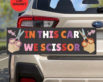 May include: A rectangular car magnet with a dark gray background featuring the text IN THIS CAR WE SCISSOR in multicolored block letters. The design includes illustrations of two hands holding scissors, one on each side, and small pink hearts.