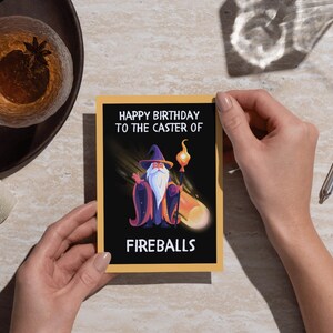 May include: A birthday card with a black background and a yellow border. The card features a cartoon wizard with a staff and the text "HAPPY BIRTHDAY TO THE CASTER OF FIREBALLS". The card is held by a person.