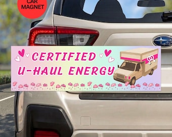 May include: A rectangular car magnet featuring a pastel gradient background with the pink text CERTIFIED U-HAUL ENERGY. The design includes pink hearts, a row of small pink flowers, and an illustration of a beige moving truck with a pink stripe.