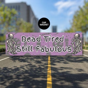 May include: A rectangular car magnet with the text "Dead Tired Still Fabulous" in white, outlined in black, on a purple background. The magnet features cartoon skeletons on each end and a black circle with "CAR MAGNET" in white.
