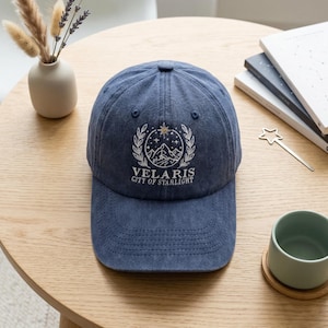 May include: A denim blue baseball cap with the text "VELARIS CITY OF STARLIGHT" embroidered in white. The design includes a mountain scene, stars, and laurel leaves. The cap is sitting on a wooden table.