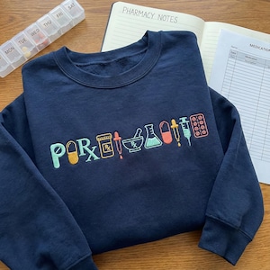 Peut inclure: Sweat-shirt bleu marine avec des graphiques brodés sur le thème de la pharmacie. Le motif comprend le mot "POR" et divers symboles médicaux multicolores. Un pilulier et un carnet intitulé "PHARMACY NOTES" sont également visibles.