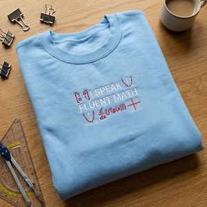 May include: A light blue sweatshirt with the embroidered text "I SPEAK FLUENT MATH" in white and red. The design includes mathematical symbols and equations. The sweatshirt is folded on a wooden surface.