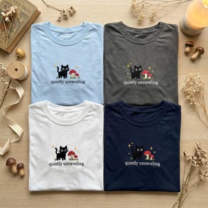 May include: Four t-shirts in light blue, grey, white, and navy blue, each with an embroidered design. The design features a black cat, red and white mushrooms, and the text "quietly unraveling".