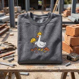 Könnte beinhalten: Dunkelgraues Sweatshirt mit Rundhalsausschnitt und einer Cartoon-Gans, die einen gelben Bauhelm trägt und einen weiteren hält. Die Gans ist von Ziegeln und Werkzeugen umgeben. Ein lustiges Design für Bauarbeiter oder Gänseliebhaber.