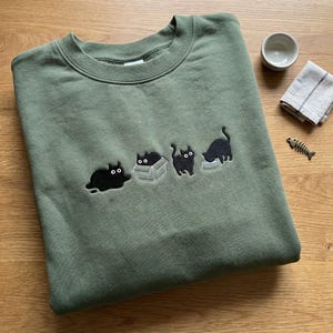 May include: Olive green sweatshirt with an embroidered design of four black cats in various poses. One cat is lying down, one is in a box, one is standing, and one is eating from a bowl. A small bowl, a napkin, and a fish-shaped decoration are in the background.