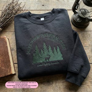 May include: Dark grey sweatshirt with a green embroidered design. The design includes the text "MIRKWOOD NATIONAL PARK", trees, and a deer, with "tread lightly traveler" below. The sweatshirt is folded on a wooden surface.