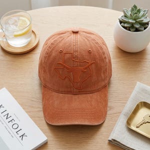 May include: A burnt orange baseball cap featuring an embroidered outline of the state of Texas with a longhorn design. The cap has a curved brim and a classic six-panel construction. A casual accessory for everyday wear.